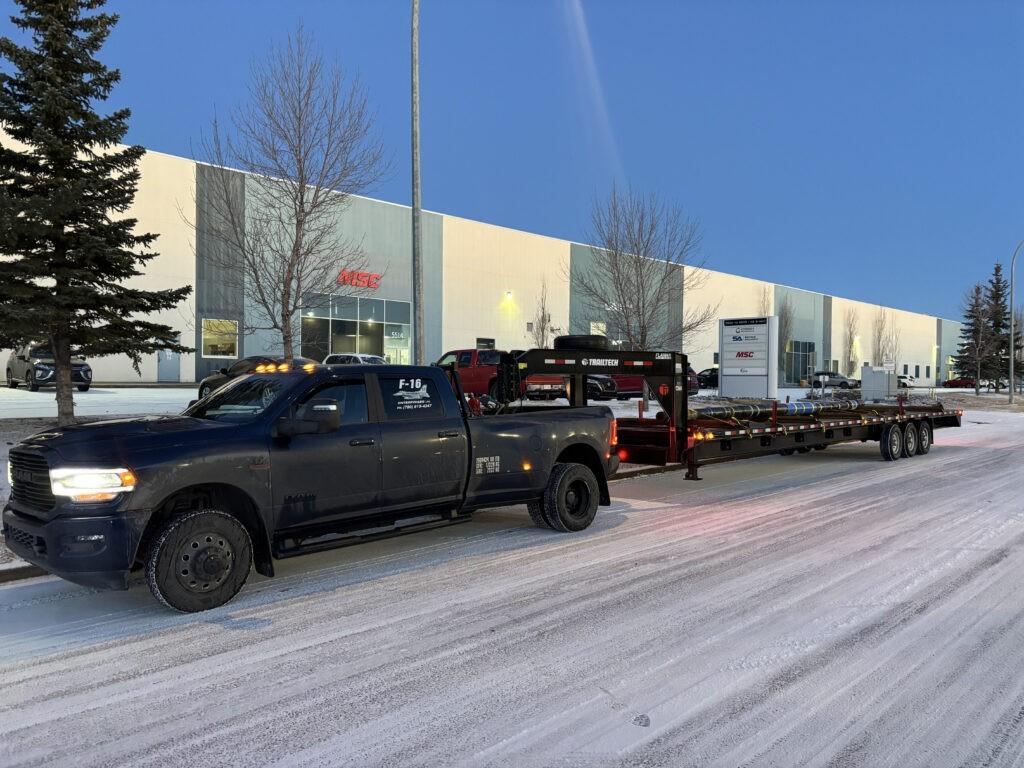 Two trucks parked on a snowy street near industrial buildings under a clear blue sky.