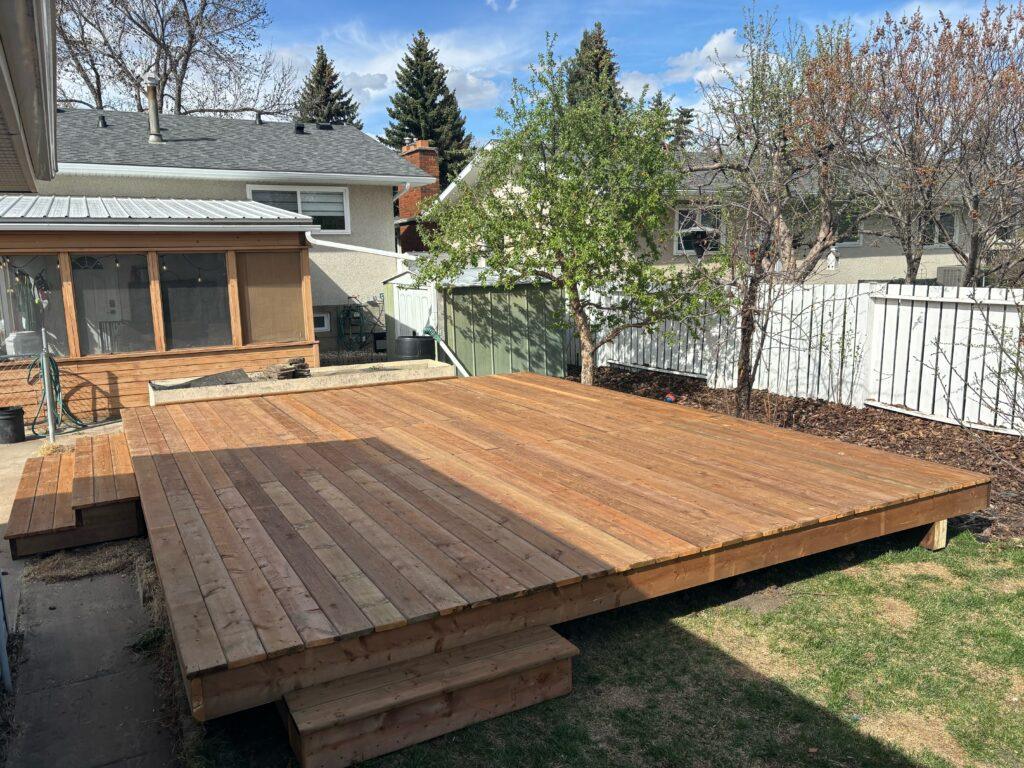 A newly built wooden deck with steps in a backyard under a clear sky.