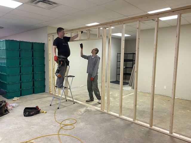 Two men building a wooden frame wall inside a room.