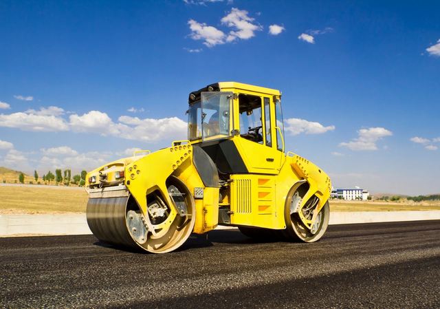 Bright yellow road roller compacting asphalt under a blue sky.