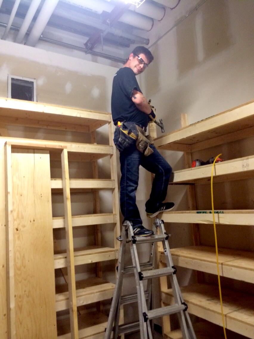 Man standing on a ladder installing wooden shelves in a room.