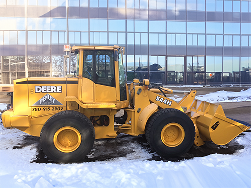 Yellow John Deere wheel loader parked on snow near a glass building.