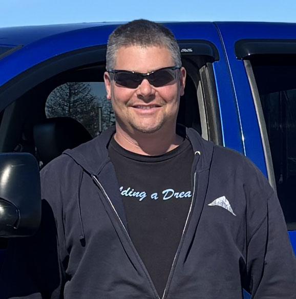 Man wearing sunglasses and a black hoodie stands by a blue vehicle.