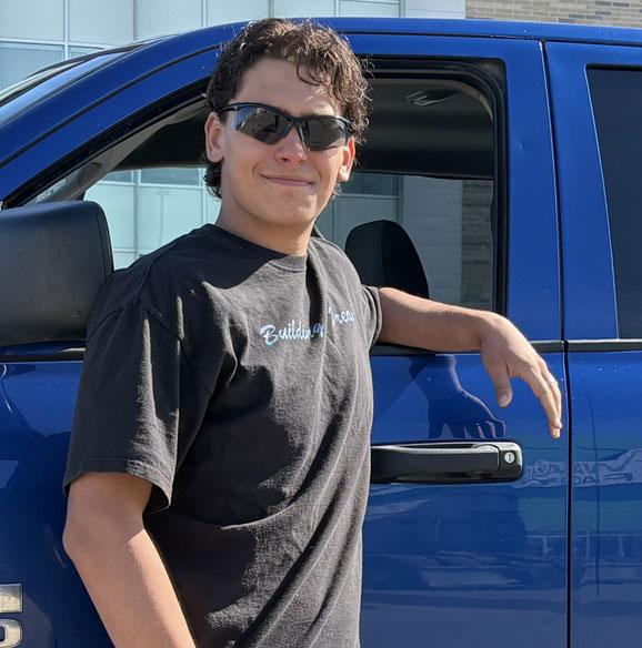A young man in sunglasses leans casually on a blue truck.